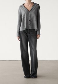Persona che indossa una camicia grigia a righe con collo ampio e jeans neri larghi, in piedi su un pavimento semplice contro un muro bianco.