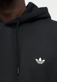 Gros plan d'une personne portant un sweat à capuche noir avec un logo blanc Adidas trefoil sur la poitrine gauche et des cordons noirs.