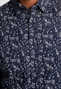 Camisa azul marino de botones con un patrón de cachemira en blanco y gris. Hecha de tela tejida con una textura suave y cuello clásico.