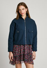Giacca corta blu navy con colletto alto, zip frontale e due ampie tasche a pattina, abbinata a un vestito con fantasia floreale.