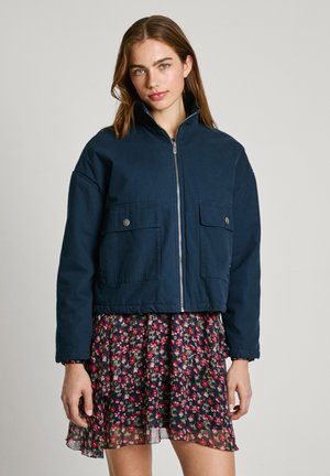 Marinblå kort jacka med hög krage, framtida dragkedja och två stora ficklock, tillsammans med en klänning med blommönster.