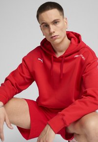Piros kapucnis pulóver zsinórral és logóval, piros nadrággal párosítva. Puha anyaggal és laza szabással rendelkezik. Egyszerű, sportos dizájn, minta nélkül.