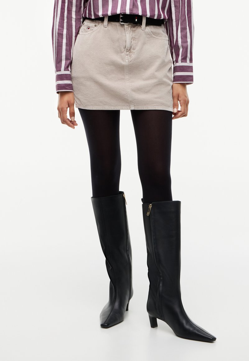 Femme portant une mini-jupe en velours côtelé beige, des collants noirs, des bottes noires montantes avec des fermetures éclair, et une chemise rayée violette et blanche.