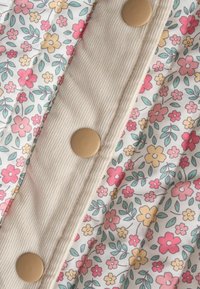 Close-up van beige ribfluwelen stof met grote ronde knopen naast witte stof met een patroon van kleine roze, gele en koraalrode bloemen en groene bladeren.