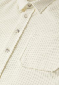 Gros plan sur une chemise en tissu côtelé crème avec des boutons beiges, un col et une poche poitrine à droite.