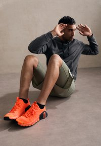 Lyse orange sportsko med sorte detaljer, der har en tekstureret overflade og fleksibel sål, parret med en grå hoodie og olivengrønne shorts.