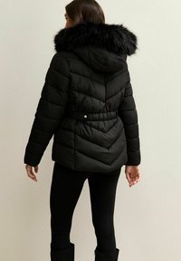 Kobieta w czarnej pikowanej kurtce puchowej z kapturem i czarnym sztucznym futrem, w połączeniu z czarnymi legginsami i butami, stojąca tyłem.