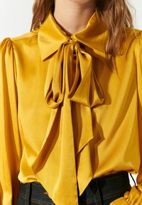 Blusa di raso gialla con un grande fiocco legato al collo, maniche plissè e texture liscia, abbinata a pantaloni in denim scuri a vita alta.