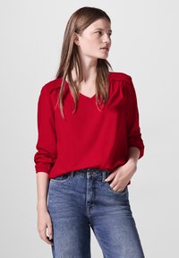 Rode blouse met een V-hals, gerimpelde schouderdetails en lange, iets verzamelde mouwen. Gepaarde met blauwe denim jeans. Gladde stofstructuur.
