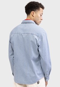 Light denim shirt with vertical white stripes, point collar, button cuffs, and a logo patch on the lower hem. Soft texture and relaxed fit.