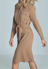 Abito camicia color tan con maniche lunghe, colletto e doppie tasche sul petto. Presenta una vita annodata e una texture liscia. Lunghezza a metà polpaccio.