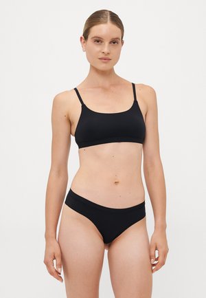 Vrouw met lichte huid en bruin haar dat teruggebonden is, gekleed in een zwarte sportbeha en bijpassende zwarte bikini-onderdelen, staande tegen een witte achtergrond.