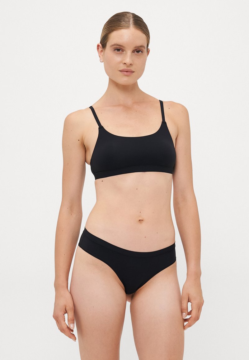 Woman with light skin and brown hair tied back, wearing a black sports bra and matching black bikini bottoms, standing against a white background.