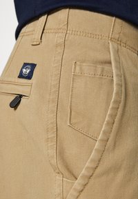 Pantalon en coton beige avec une poche zippée et un patch tissé bleu portant un logo de lion. Coutures visibles et texture lisse.