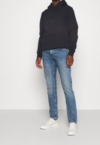 Sudadera azul marino con bolsillo rectangular y cordones, combinada con jeans de mezclilla azul claro y zapatillas blancas. Diseño simple y casual.