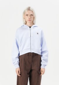 Sudadera recortada de color azul claro con cremallera en el frente, capucha con cordón y detalle de parche. Combinada con pantalones cargo marrones. Textura de tela suave.
