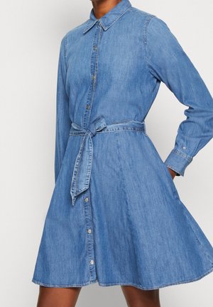 Denimskjortekjole med lange ærmer, knapper foran, krave og bundet bælte i taljen, båret af en person med den ene hånd i lommen.