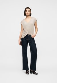 Γυναίκα που φοράει μπεζ τοπ με V-λαιμόκοψη, σκούρο τζιν με φαρδιά μπατζάκια και μαύρα παπούτσια με τακούνι, στέκεται με το ένα χέρι στην τσέπη και κρατά ένα τσαντάκι.