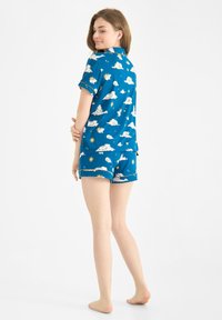 Kortärmad blå pyjamasset med mönster av vita moln och sol. Har en kragarskjorta och matchande shorts, tillverkade av mjukt tyg.