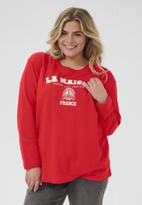 T-shirt rouge à manches longues avec un col rond, arborant le texte blanc "LA MAISON" et des graphiques. Tissu doux avec une coupe décontractée.