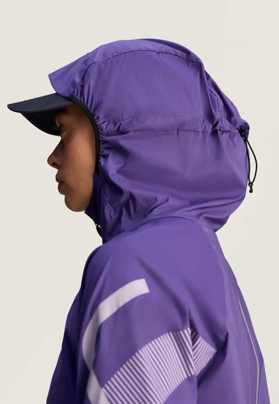 Chaqueta con capucha impermeable de color púrpura con un acabado texturizado, que presenta una visera negra y acentos gráficos blancos. Vista de perfil lateral.