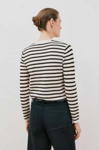 Gestreifter, langärmliger Pullover in Creme, Schwarz und Beige; taillierte Passform. Kombiniert mit dunklen Hosen mit Gesäßtaschen. Ordentlicher, glatter Stoff.