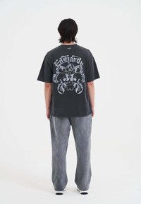 Donkergrijze katoenen t-shirt met grote witte grafische afbeelding van verstrengelde slangen en tekst op de achterkant. Gecombineerd met grijze wijde broek.