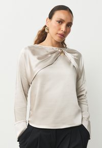 Top de satén beige con un frente drapeado y detalle de nudo en el escote, con mangas largas. La tela tiene una textura suave y brillante.