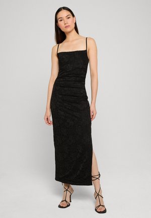 Femme portant une longue robe noire en dentelle sans manches avec bretelles fines et fente latérale, associée à des sandales noires à talons et à lanières.