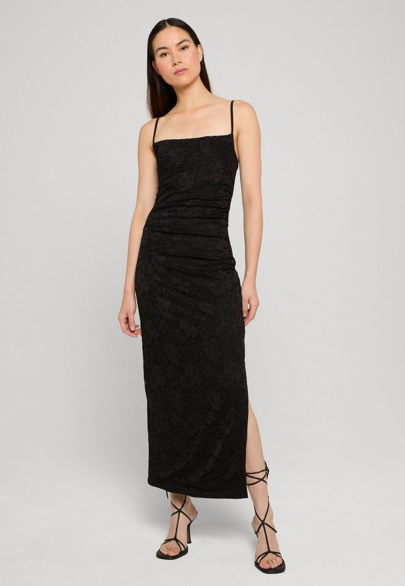 Femme portant une longue robe noire en dentelle sans manches avec bretelles fines et fente latérale, associée à des sandales noires à talons et à lanières.