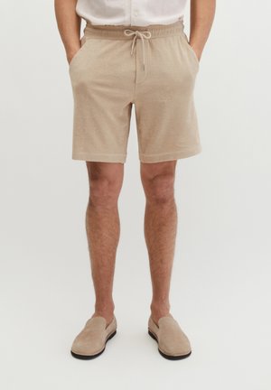 Man draagt beige shorts met een trekkoord en zijzakken, en bijpassende instapschoenen, staat met handen in de zakken.