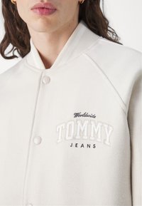 Béžová bombera z textúrovaného materiálu, s rebrovaným golierom, patentnými gombíkmi a vyšitým logom "TOMMY JEANS".