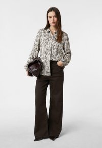 Woman wearing a snakeskin-patterned shirt, dark wide-leg pants, holding a large dark leather clutch bag with one hand in pocket.