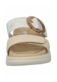 Sandales beige et argentées avec un orteil arrondi, dotées d'une large sangle de cheville et d'un accent circulaire marron. Matériaux en daim et texturés.