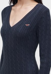 Pull bleu marine en maille torsadée avec col en V et petit logo oiseau rouge et blanc sur la poitrine, porté par une personne des épaules à la taille.