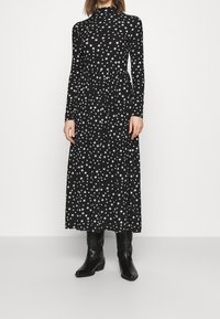 Svart långärmad klänning med hög halsringning, prydd med ett vitt stjärnmönster, flödande silhuett och bärs med svarta cowboyboots.