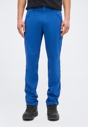 Muž v jasne modrých vonkajších nohaviciach a čiernych turistických topánkach stojaci na jednotnom pozadí.