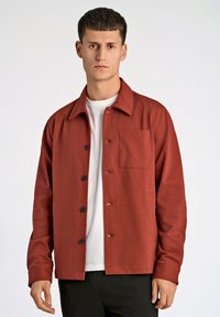 Rote Hemdjacke mit Knopfleiste, Kragen und einer Brusttasche. Hergestellt aus einem glatten Stoff mit leichter Textur. Getragen über einem weißen T-Shirt.