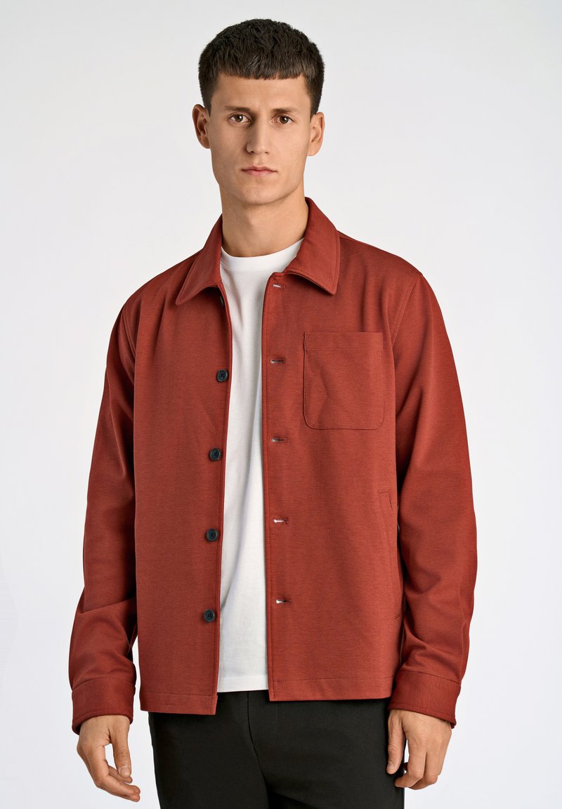Rote Hemdjacke mit Knopfleiste, Kragen und einer Brusttasche. Hergestellt aus einem glatten Stoff mit leichter Textur. Getragen über einem weißen T-Shirt.