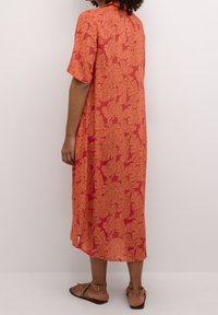 Maxi-jurk in oranje met een bladpatroon, korte mouwen, zijsplitten en een ontspannen pasvorm. Gemaakt van lichte stof. Bruine sandalen gedragen.