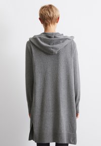Szara, długa bluza z kapturem, wykonana z miękkiego materiału, o luźnym fasonie, z kapturem i bocznymi rozcięciami ułatwiającymi ruch, z prążkowanymi akcentami przy dolnym wykończeniu.