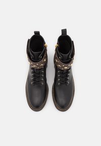 Czarne skórzane botki za kostkę z brązowym teksturowanym mankietem, ozdobione złotymi geometrycznymi ćwiekami i dekoracyjnym złotym zamkiem błyskawicznym. Okrągły nosek i sznurowany design.