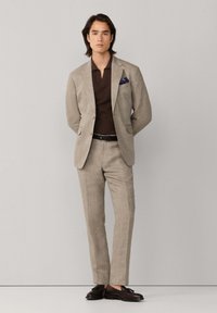 Homme debout, les mains dans le dos, portant un costume beige, un polo marron foncé, une ceinture noire et des chaussures de ville marron foncé, sur un fond uni.