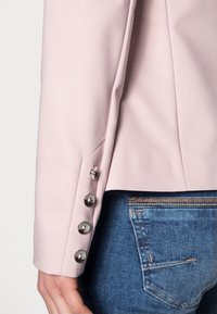 Gros plan sur une personne portant un blazer rose pâle avec quatre boutons métalliques sur la manche et un jean bleu avec des coutures de poche arrière visibles.