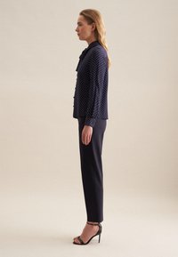 Blusa navy con pois bianchi, colletto arricciato e dettagli con bottoni, abbinata a pantaloni neri aderenti e tacchi neri con cinturini.
