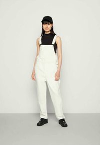 Jovem mulher vestindo um macacão branco, blusa preta sem mangas, chapéu preto e ténis pretos, de pé diante de um fundo claro e simples.