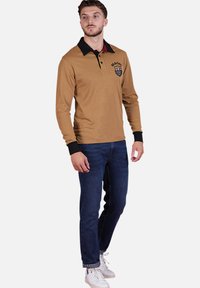 Polo à manches longues couleur beige avec un col et des poignets noirs, comportant un patch logo. Associé à un jean bleu foncé et des baskets blanches.