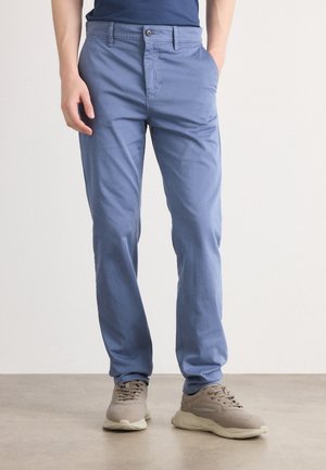 Mann iført lyseblå casual bukser og beige sneakers, stående med den ene hånden i lomma på en ensfarget bakgrunn.