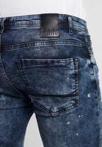 Jeans de mezclilla en un lavado azul desgastado con un parche de cuero etiquetado "CARS", con dos bolsillos traseros y detalles desgastados.