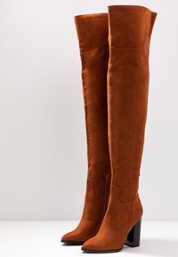 Bottes hautes en daim marron jusqu'au genou avec un bout pointu, un talon bloc et un détail de fermeture éclair sur le côté. Texture lisse et design ajusté.